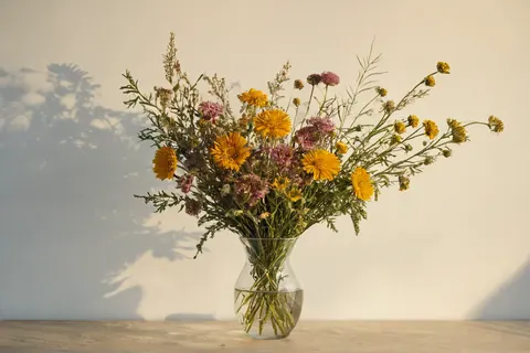A glass vase with yellow and pink flowers on a wooden table against a white wall with shadows.