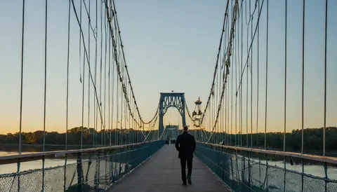 A man in a dark suit walks away on a wooden suspension bridge over a river at sunset.