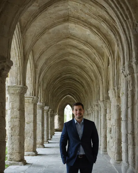 A man in a navy suit smiles standing in a historic stone cloister with pointed arches.