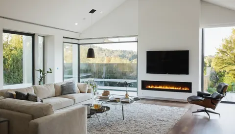 A modern living room with beige sofa glass table fireplace and large windows overlooking greenery