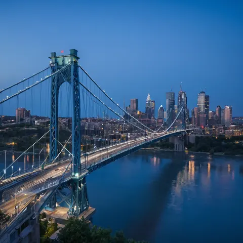 A suspension bridge over a river with a city skyline lit up at twilight.