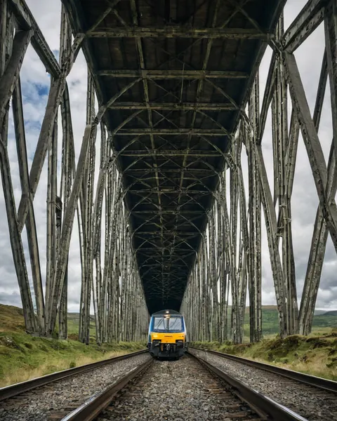 A train on a railway track under a large steel viaduct with green hills in the background.