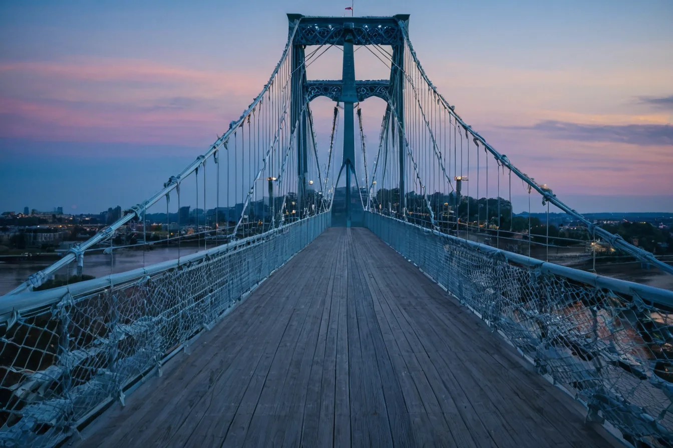 A wooden pedestrian bridge with metal railings at twilight over a river with city lights in the distance.