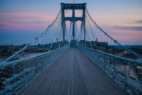 A wooden pedestrian bridge with metal railings at twilight over a river with city lights in the distance.