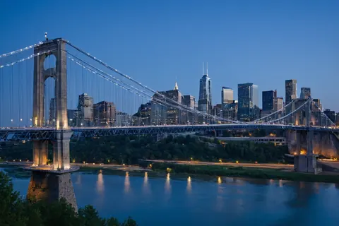 Brooklyn Bridge lit at dusk over river with Manhattan skyline in background