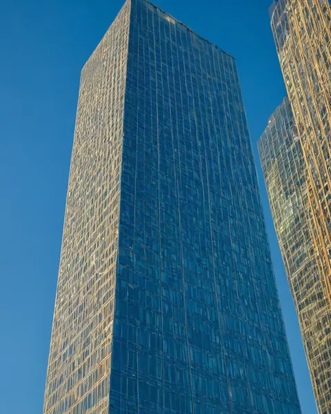 Towering glass skyscraper with blue reflective facade and golden sunlit edges against clear blue sky