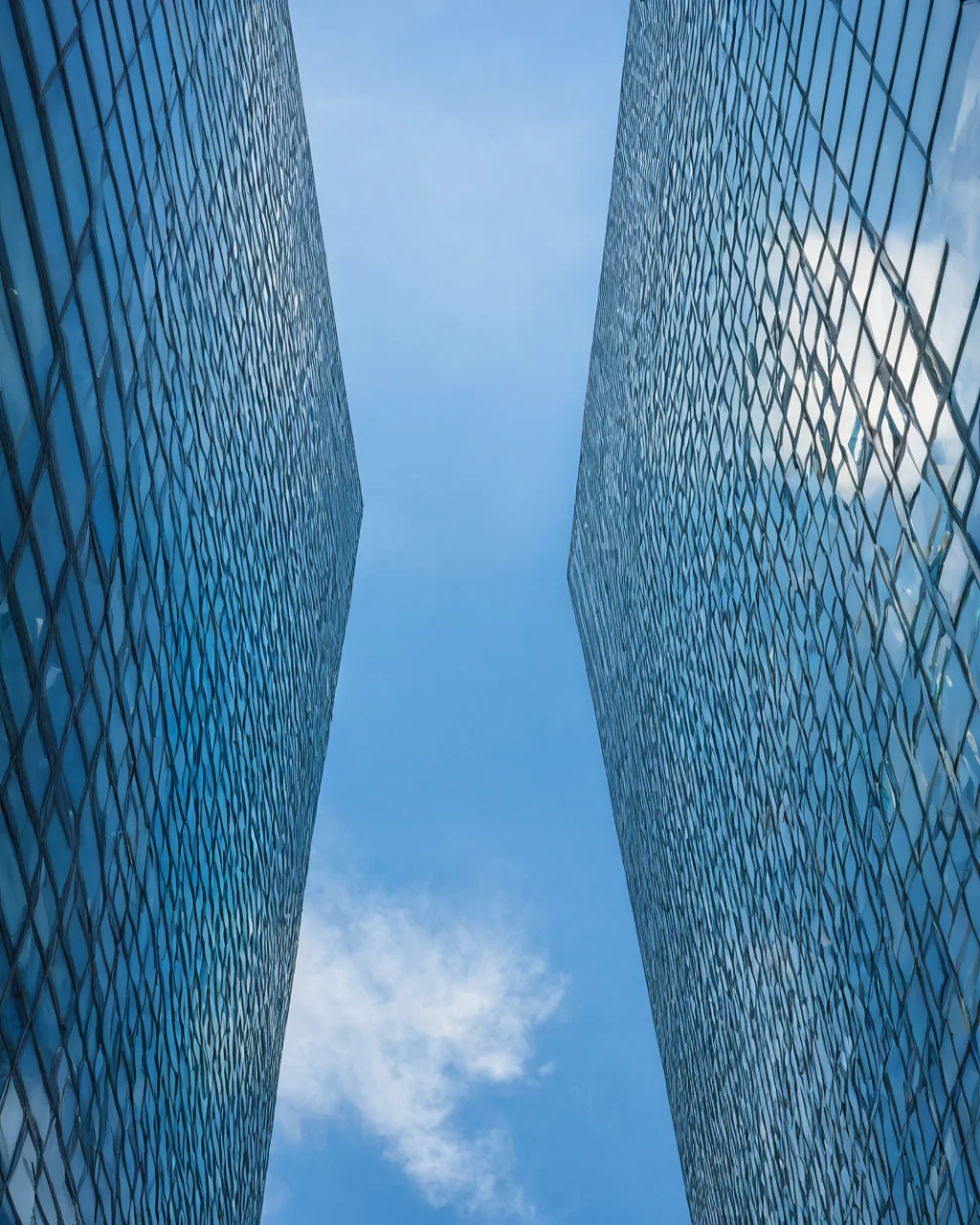 Two modern glass skyscrapers with diamond patterns frame a blue sky with white clouds.
