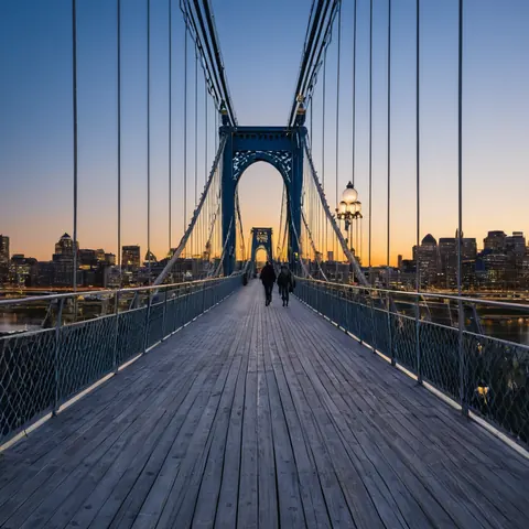 Two people walking on a wooden suspension bridge toward a city skyline at dusk.