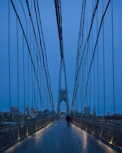 Two people walking on a wooden suspension bridge toward a city skyline at dusk.