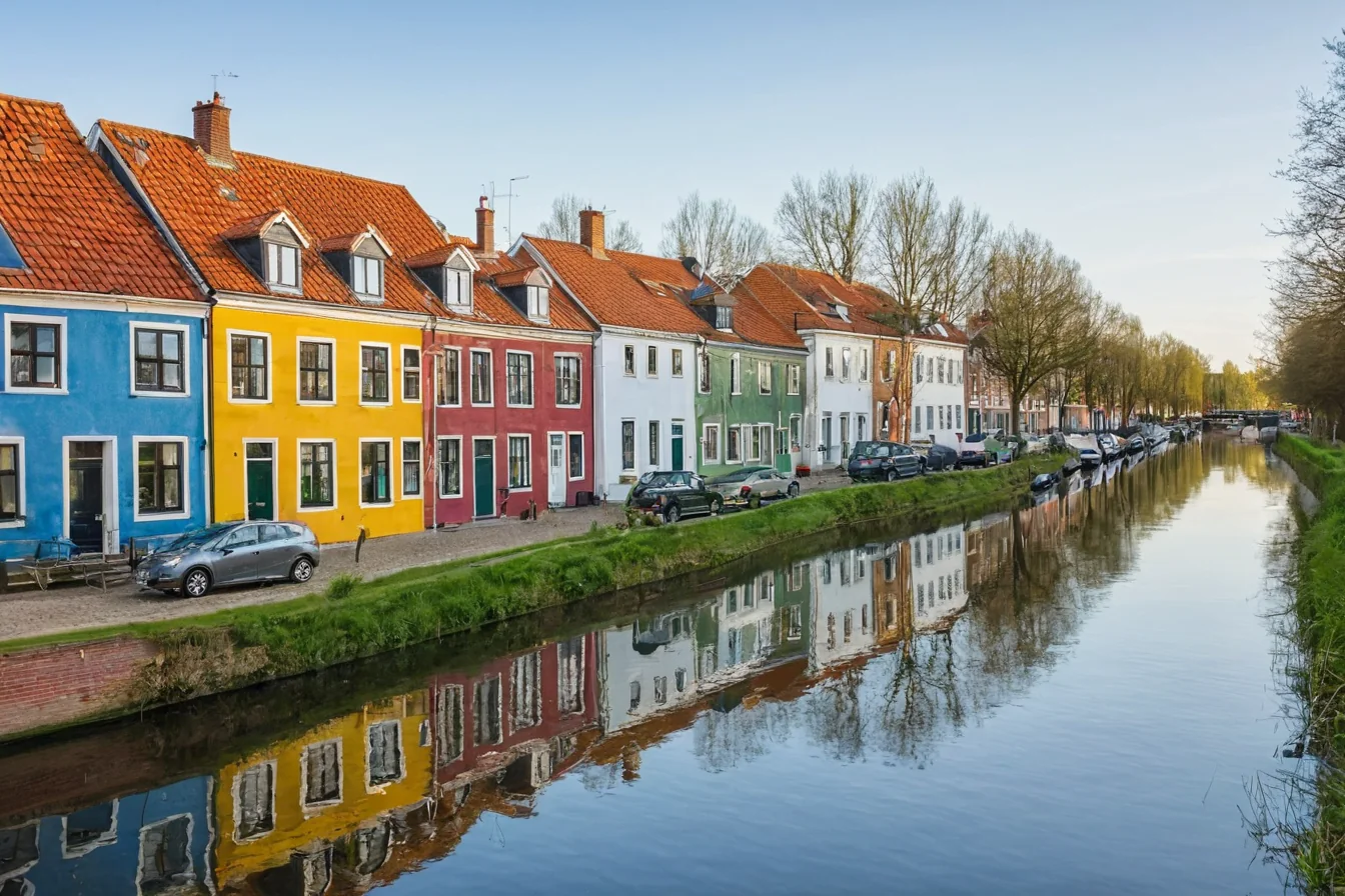 Vibrant row houses with red roofs line a calm canal reflecting their colorful facades and bare trees under a clear sky.